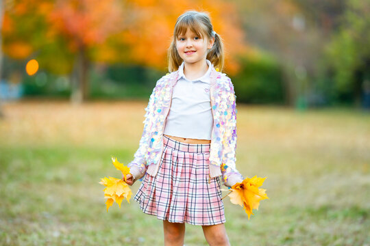 Child girl holds yellow leaves. Child wears sparkling jacket. Smiling child girl stands in park. Cute child portrait outdoors.