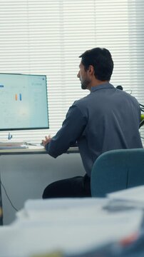 Vertical Screen : Rear view young Latin businessman analyzing financial graph data on computer screen in office with paperwork. Hybrid work, fintech strategy, corporate performance, market statistics.