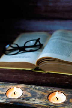 Open book with reading glasses resting on pages, illuminated by two warm glowing candles, representing knowledge and cozy reading