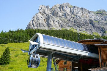 Modern mountain cable car station with glass gondola cabin beside alpine lodge, set below rugged rocky peak, green forest slope, clear blue sky, bright summer daylight, scenic transport access © DIgnat_Switzerland