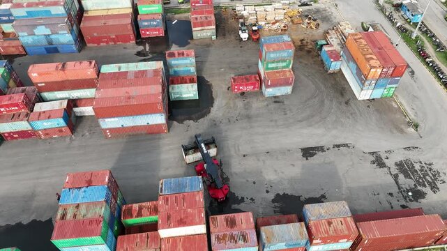 High-angle aerial drone shot of a busy logistics terminal featuring a reach stacker crane moving industrial shipping containers in a large storage yard for global import export trade operations.