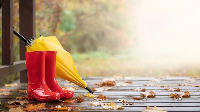 Red kids rubber boots and yellow umbrella on porch with blurred autumn garden background banner.