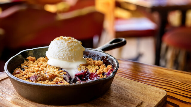 Baked plum crumble with vanilla ice cream in skillet with blurred restaurant background banner.