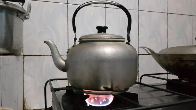 Aluminum kettle being heated over a visible gas flame on a stove in a simple home kitchen with tiled walls..