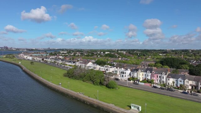 Colorful terraced houses line Clontarf&rsquo;s bayfront as a green park and seafront road stretch along the calm water under blue skies.