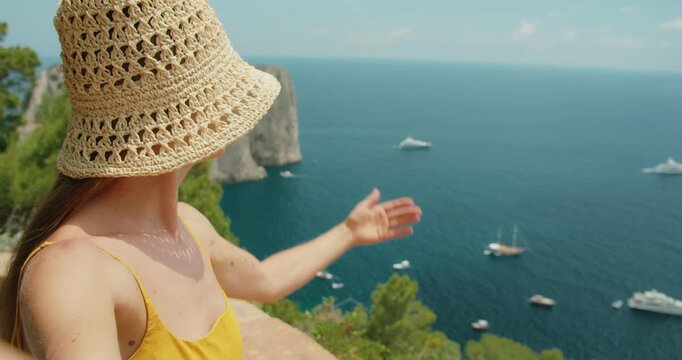 Smiling woman in straw hat recording selfie video showing Faraglioni sea stacks above deep blue Capri waters on sunny summer day