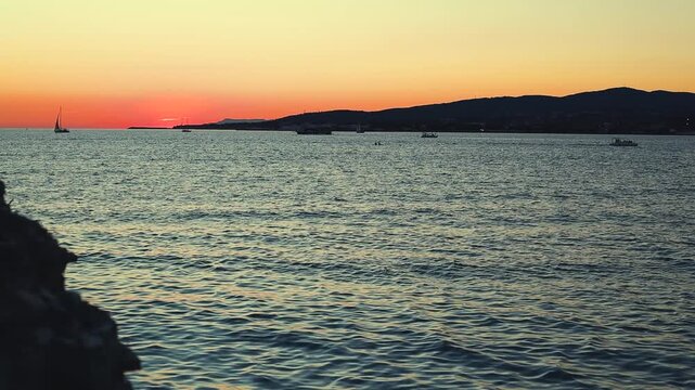 Vibrant sunset casts orange and purple hues over calm sea, with silhouetted boats and distant hills creating a tranquil coastal scene
