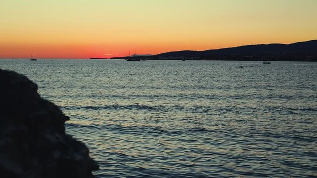 Vibrant sunset casts orange and purple hues over calm sea, with silhouetted boats and distant hills creating a tranquil coastal scene