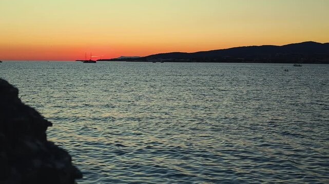 Vibrant sunset casts orange and purple hues over calm sea, with silhouetted boats and distant hills creating a tranquil coastal scene