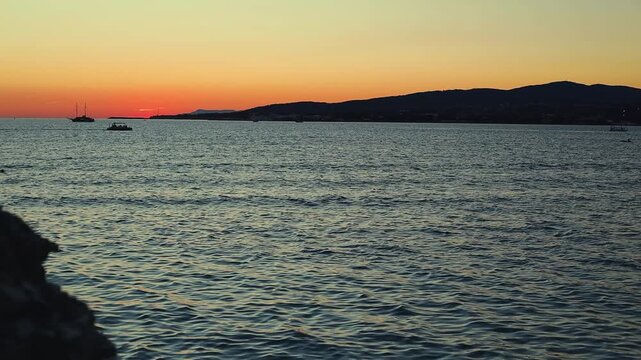 Vibrant sunset casts orange and purple hues over calm sea, with silhouetted boats and distant hills creating a tranquil coastal scene