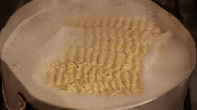 Close-up (zoom out) of instant noodles cooking in a pot. Noodles boiling in a pot on the stove. Pasta cooking in a pot of hot water. POV: making instant ramen in the kitchen; preparing macaroni. 