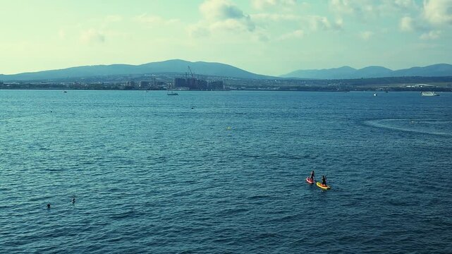Two paddleboarders navigate calm waters, surrounded by distant hills and boats, highlighting a peaceful coastal scene under a clear sky