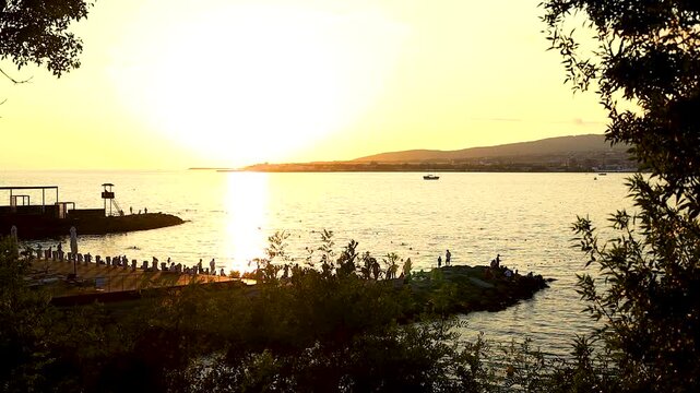 Sunset casts a warm glow over the water, highlighting silhouettes of people along the shore and boats in the distance, framed by greenery