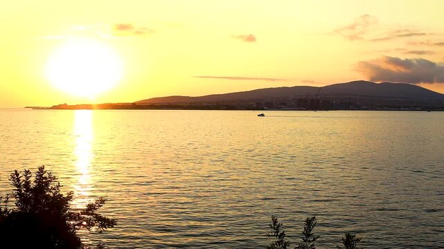 Sunset casts a warm glow over the water, highlighting silhouettes of people along the shore and boats in the distance, framed by greenery