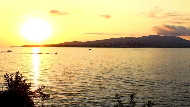 Sunset casts a warm glow over the water, highlighting silhouettes of people along the shore and boats in the distance, framed by greenery
