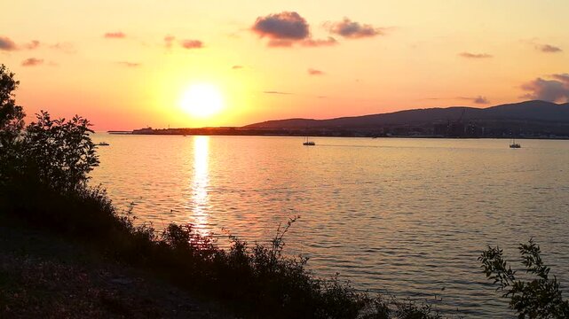 Sunset casts warm hues over water as boats sail peacefully, framed by trees and distant hills, creating a tranquil coastal atmosphere