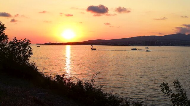 Sunset casts warm hues over water as boats sail peacefully, framed by trees and distant hills, creating a tranquil coastal atmosphere