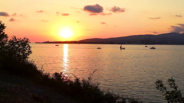 Sunset casts warm hues over water as boats sail peacefully, framed by trees and distant hills, creating a tranquil coastal atmosphere