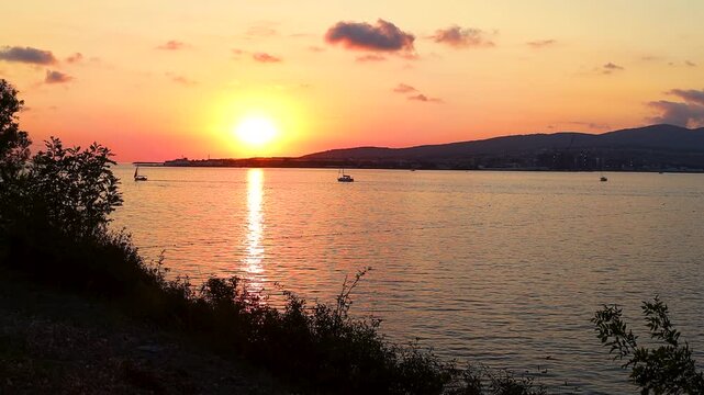Sunset casts warm hues over water as boats sail peacefully, framed by trees and distant hills, creating a tranquil coastal atmosphere