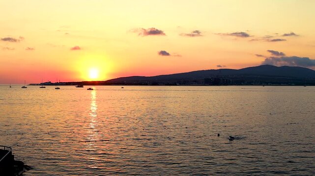 Sunset casts vibrant orange and yellow colors over the ocean, with boats gently bobbing on the water's surface, creating a serene evening atmosphere