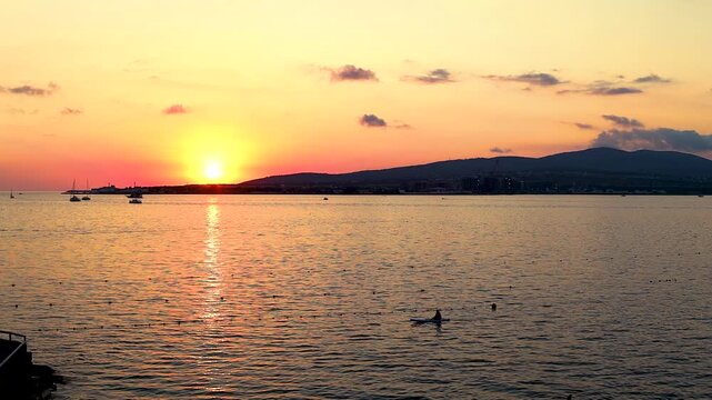 Sunset casts vibrant orange and yellow colors over the ocean, with boats gently bobbing on the water's surface, creating a serene evening atmosphere
