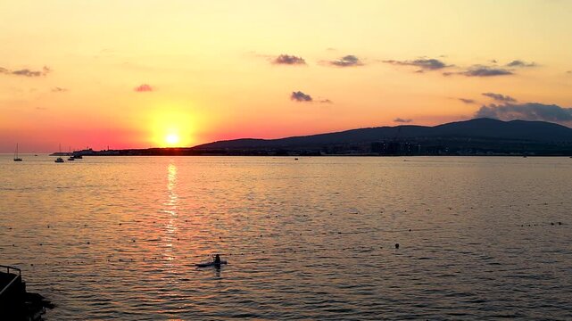 Sunset casts vibrant orange and yellow colors over the ocean, with boats gently bobbing on the water's surface, creating a serene evening atmosphere