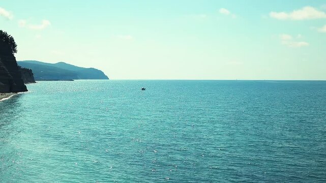 Small fishing boat glides through calm blue waters near a rocky coastline, with distant mountains and a clear sky enhancing the peaceful coastal landscape