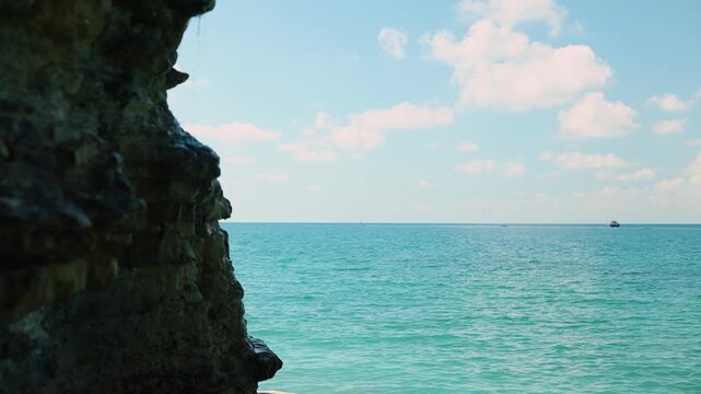 Coastal scene featuring turquoise ocean waves meeting rocky shore under a clear blue sky with fluffy clouds and a distant boat on the horizon
