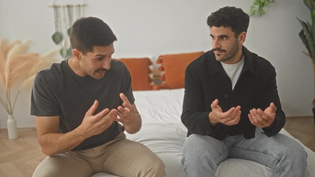 Two men sit on a bed in a building, hands gesturing while one speaks and the other listens, visible mustache and jacket details; tension friendship.