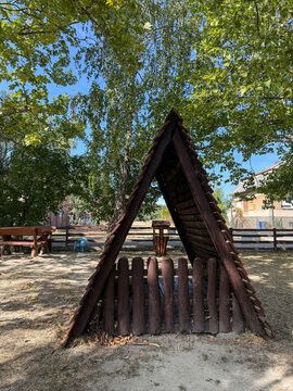 A small triangular dark wooden playhouse standing next to a fence on a children playground