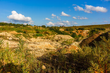 Obraz premium Rugged terrain of an abandoned sandy quarry with dry vegetation and loose rocks under a bright blue summer sky with fluffy white clouds on a sunny day in the countryside