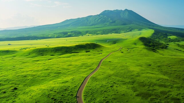 A vast meadow of fresh greenery and a winding road stretching out against a backdrop of blue skies and majestic mountain ranges (drone aerial shot)