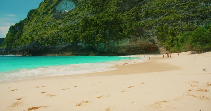 Beachgoers enjoying sunny day on white sand beach at base of steep green cliff in Nusa Penida Bali