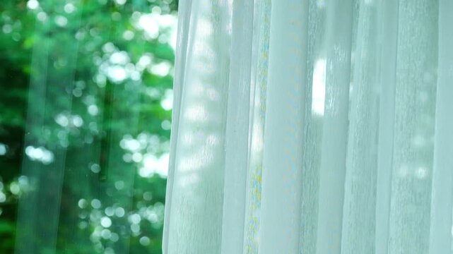 Footage of white sheer curtains covering a window with a soft green bokeh view of nature outside, representing a peaceful, calm, and cozy home interior atmosphere.concept home relax	