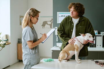 Asian female veterinarian taking notes during consultation with young adult Caucasian male pet...