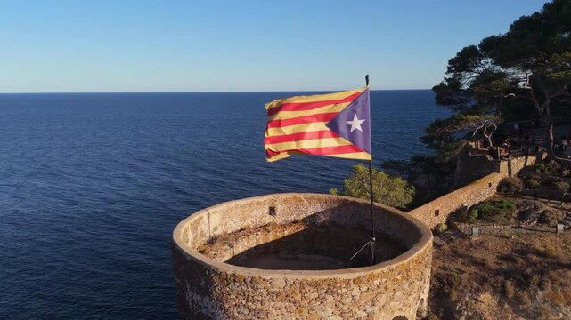The flag of Catalonia flies on the tower of a fortress on the seashore at sunset on a sunny day in the tourist town of Tossa De Mar, Catalonia, Spain