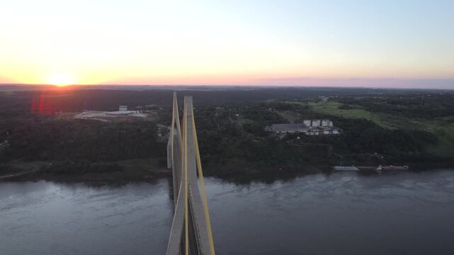 4K drone aerial view of the Integration Bridge at sunset, connecting Paraguay and Brazil across the Paran&aacute; River near the Triple Frontier. Warm evening light illuminates the bridge and surrounding lan