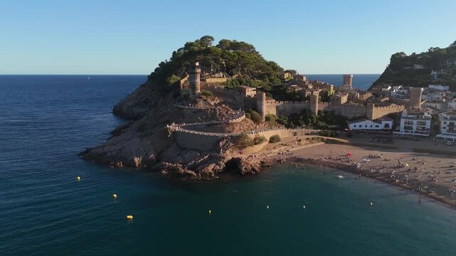 Aerial drone view of a fortress on coastal cliffs, a beach and the sea at sunset in the tourist town of Tossa de Mar, Catalonia, Spain