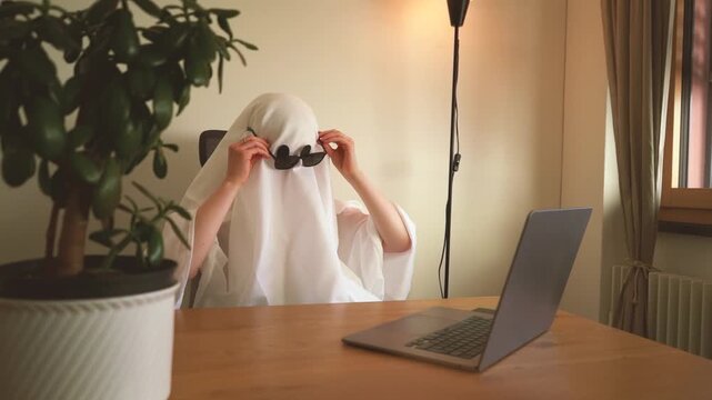 White bed sheet ghost putting on sunglasses while sitting behind a laptop in a cozy home office, creating a funny spooky remote work concept