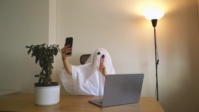 White sheet ghost taking selfies with a phone while working from home. looking cute.