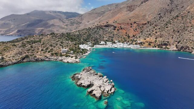 A wide aerial view reveals a pale rocky islet in turquoise water before Loutro village on Crete, backed by dry mountain slopes as a motorboat crosses the bay.