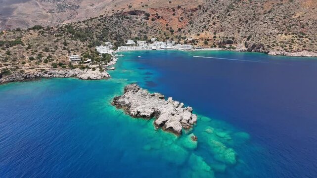Loutro Bay opens toward a turquoise cove with rugged mountains, rocky outcrops and white waterfront buildings, while a small boat approaches in the distance, drone forward flyover
