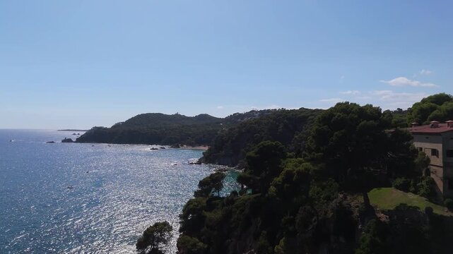 An aerial view of a drone flies from Santa Clotilde Park over the beach and coastal cliffs on a sunny day in the tourist town of Lloret De Mar, Catalonia, Spain.