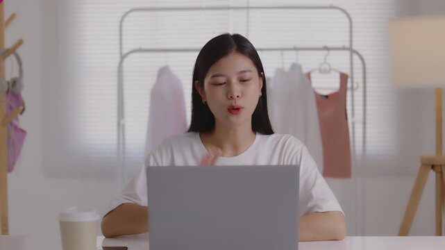 Smiling young Asian woman gesturing during a video call or online meeting on her laptop at her studio desk, representing remote work, online business, and digital communication.