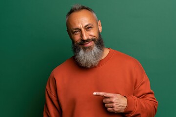 Fototapeta premium Gray bearded man pointing to the side in orange sweatshirt studio portrait on green background for lifestyle headshot and product endorsement