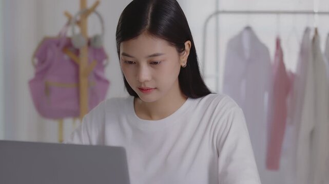 Focused young Asian woman typing on her laptop at a desk in a studio or home office, managing online business, e-commerce, or remote work.