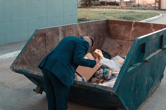 an impoverished businessman rummages in a trash can in search of food. A homeless homeless man in a suit holds burger in his hand while standing in a landfill. bankruptcy, no money concept