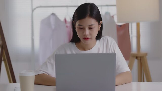 Focused young Asian woman typing on her laptop at a desk in a studio or home office, managing online business, e-commerce, or remote work.
