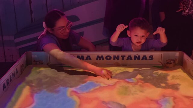 A loving mother and her two-year-year-old Latino son interact with an educational sand projection exhibit at an aquarium. The boy playfully tries to catch a whale projection, learning about beach cons
