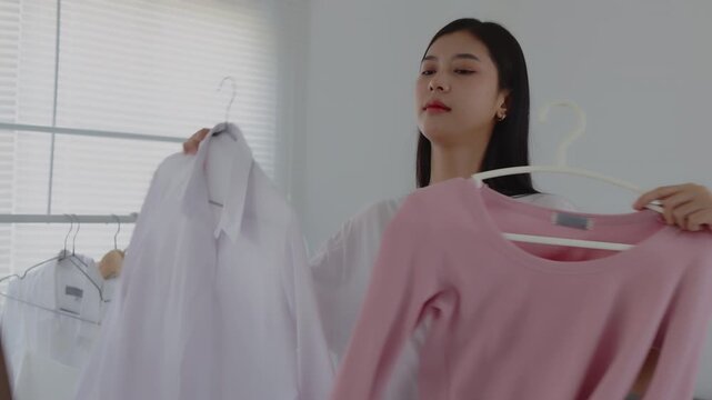 Young Asian woman holding two shirts on hangers in front of a mirror, comparing options and deciding what to wear or selecting clothes for online sale.
